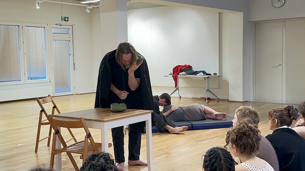 An actor speaks into a dark green rotary phone that is on a table while two other actors are behind him in mid-performance of 'Faith Hope Charity'. One actor is lying down with their back to the audience while the third actor is seated with his forehead resting on the second actor's shoulder. Members of the audience are seen on the right side of the photo, watching the performance. 